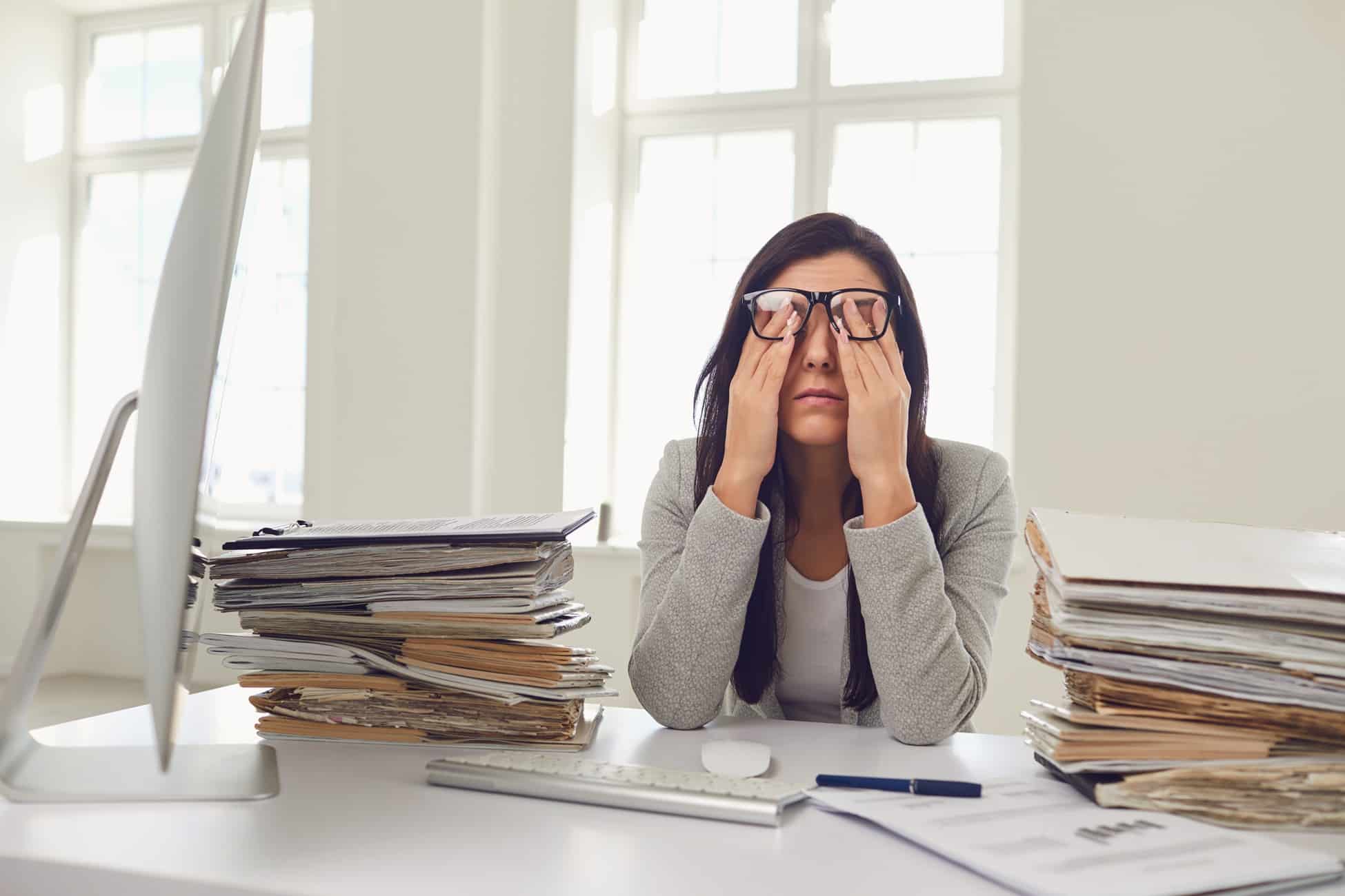 Overwhelmed professional woman rubbing eyes surrounded by stacks of paperwork