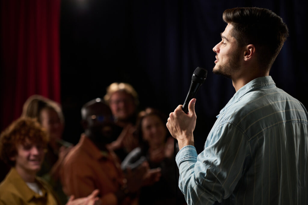 Man performing with microphone on stage in front of diverse audience seen clapping and enjoying the performance