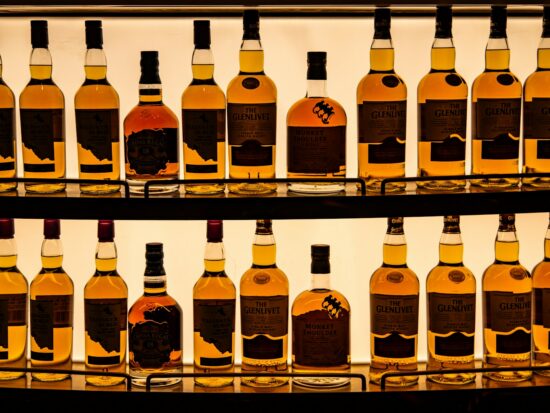 A display of whiskey bottles arranged on backlit shelves. The collection includes brands such as The Glenlivet, Talisker, Chivas Regal, and Monkey Shoulder. The amber liquid in the bottles is illuminated by the light from behind, highlighting the labels and creating a warm, inviting ambiance. The bottles are neatly organized, showcasing a variety of premium and well-known whiskey brands.