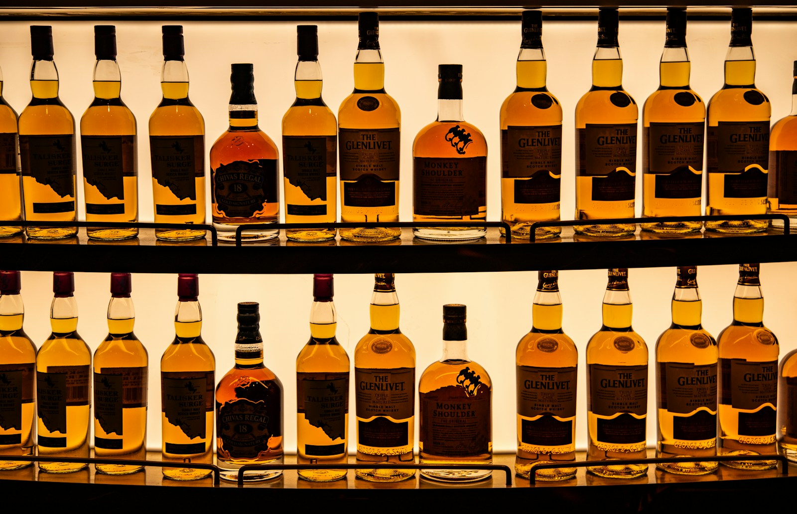 A display of whiskey bottles arranged on backlit shelves. The collection includes brands such as The Glenlivet, Talisker, Chivas Regal, and Monkey Shoulder. The amber liquid in the bottles is illuminated by the light from behind, highlighting the labels and creating a warm, inviting ambiance. The bottles are neatly organized, showcasing a variety of premium and well-known whiskey brands.